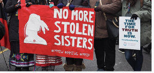 Hands encircling a large red banner with white letters reading "No More Stolen Sisters" and a smaller sign reading "The Time for Truth is Now," from the 2019 Women's March on Washington  
