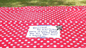 A close up of a note on a polka-dot tablecloth on a picnic table outside. It reads "Reserved for a birthday party @ 11am Thank you for being kind & leaving this here. Thank you!" The Self Help Radio logo is on the top righthand corner of the note.